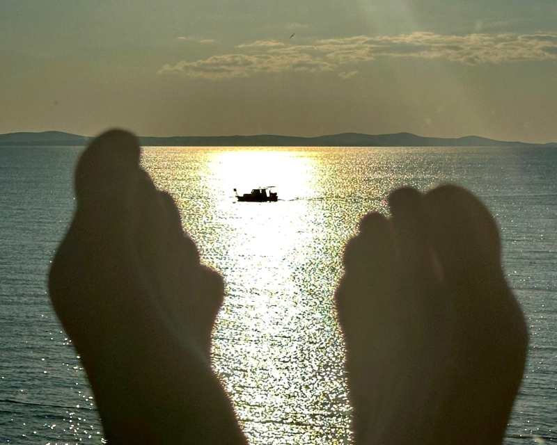 Füsse im Vordergrund beim Entspannen am Meer, im Hintergrund Boot im Sonnenlicht – Symbol für Loslassen und innere Ruhe.